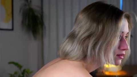 Media: Video of a woman with shoulder-length blonde hair, turned away, showing her bare back, in a dimly-lit room with green plants and abstract art on the wall.