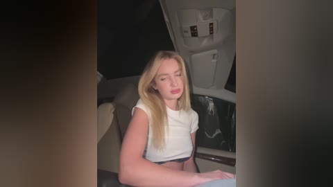 Media: Video of a young woman with long blonde hair, wearing a white crop top and black pants, sitting in a car's backseat, looking tired.