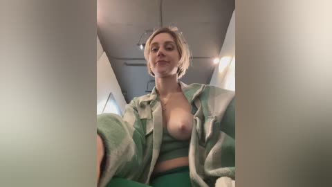 A video of a young, light-skinned woman with short blonde hair, wearing a green and white plaid shirt open to reveal her bare breasts, taken inside a dimly lit airplane cabin.