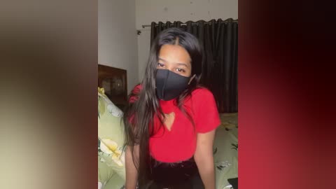 Media: Video of a young South Asian woman with long black hair, wearing a red shirt, black face mask, and sitting on a bed with floral sheets.