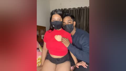 Media: A video shows a South Asian couple with light brown skin and black hair, wearing black masks and red shirts. The man has a beard and is holding the woman, who has a heart-shaped cutout on her shirt. They stand in a dimly lit room with a floral bedspread.