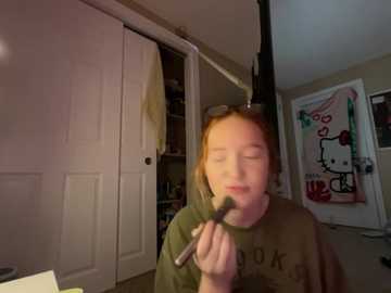 Media: Video of a young girl with red hair, wearing a green T-shirt, applying makeup in a dimly lit bedroom with white doors, a Hello Kitty poster, and shelves filled with personal items.