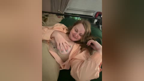 Media: Video of a smiling, fair-skinned woman with long, wavy red hair, wearing a peach robe, lying on a green bed with white blinds in the background.