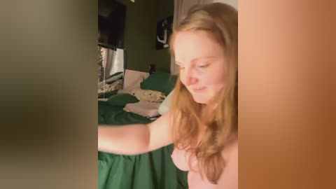 Media: A video of a young Caucasian woman with long, wavy blonde hair, wearing a pink top, sitting on a bed with green sheets. She has a gentle smile, looking at a phone in her hand. The background features a dark green wall and a TV.