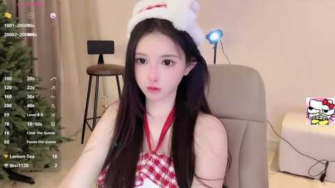 A video of a young woman with long black hair, wearing a Santa hat and red plaid top, sitting in a beige chair in a cozy living room with a Christmas tree.