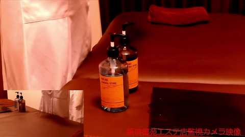 Media: Video of two clear plastic bottles labeled \"Hand Sanitizer\" on a wooden nightstand beside a neatly made bed with orange sheets, under warm lighting.