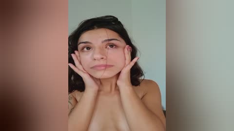 Media: A video of a young, light-skinned woman with long, dark hair, large brown eyes, and a petite physique. She is topless, resting her hands on her cheeks, gazing introspectively. The background is a neutral, off-white wall.