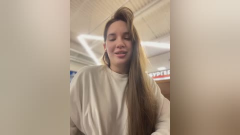 Media: Video of a smiling young woman with long, straight brown hair, wearing a white turtleneck sweater, standing indoors with a fluorescent-lit ceiling and blurred background.