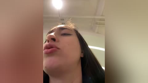 Media: Video of a young woman with straight black hair, light skin, and full lips puckered, captured from a low angle, in an indoor setting with white ceiling and beige walls.