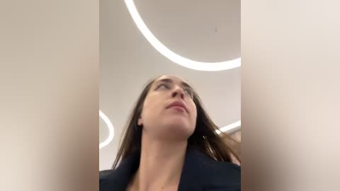 Media: A video of a young woman with straight brown hair, wearing a dark blazer, looking upwards towards the ceiling with circular LED lights.