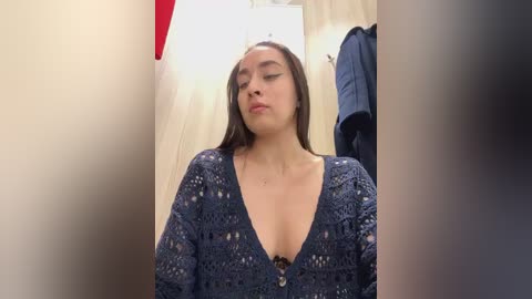 Media: A video of a young woman with long brown hair and light skin, wearing a deep V-neck, patterned blue sweater, standing in a dressing room with a red hat, coat hangers, and a mirror in the background.