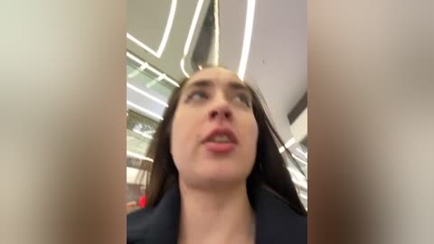 Media: Video of a young woman with long dark hair and fair skin, wearing a black jacket, captured from a low angle in a modern, brightly lit indoor space with sleek, white and gray architectural features.