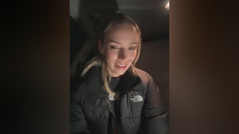 Media: A video of a blonde woman in a black puffer jacket, smiling, partially obscured by darkened car windows, inside a dimly lit car.