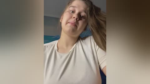 Media: Video of a young woman with light skin and straight, light brown hair, wearing a white t-shirt, leaning slightly forward with a relaxed, slightly tilted head. Background shows a blurred blue wall and indistinct furniture.