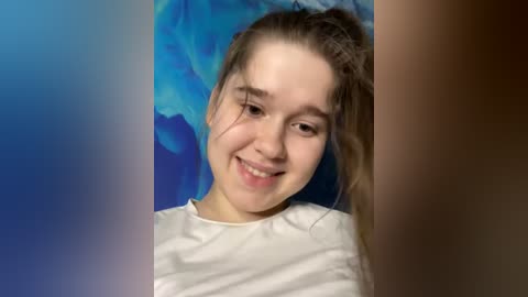 Media: Video of a smiling young girl with light skin, light brown hair, wearing a white shirt, against a blue and brown background.