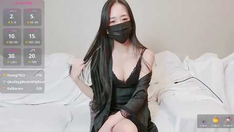 Media: Video of an Asian woman with long black hair, wearing a black mask, black lace lingerie, and robe, sitting on a white bed. Background shows a digital clock displaying 2:55 PM.