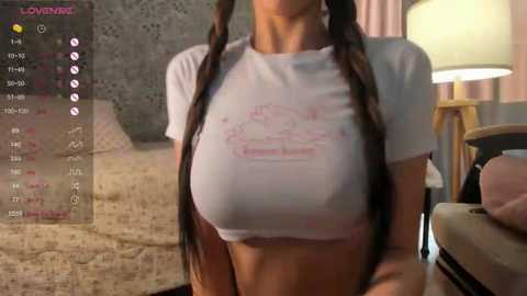 Media: A video of a woman with long brown pigtails, wearing a white crop top that reveals her large breasts, standing in a cozy bedroom with a bed, lamp, and soft lighting.