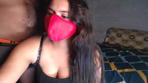 Media: A video shows a woman with long, dark hair and a red mask, wearing a black bra, in a bedroom with a blue and yellow patterned bedspread.