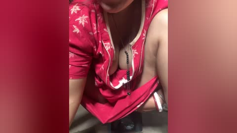 Media: Video of a woman with fair skin, wearing a red shirt with a white pattern, exposing her large breasts and cleavage, sitting in a red-tinted bathroom.