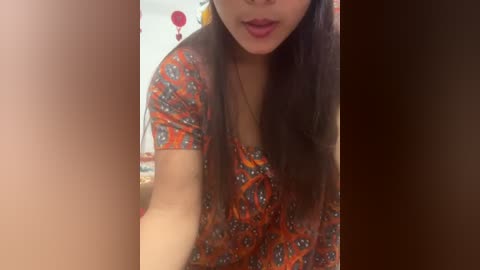 Media: Video of a young Asian woman with long brown hair, wearing a patterned orange top, partially visible. Background features a blurred, indoor setting with red and white decor.