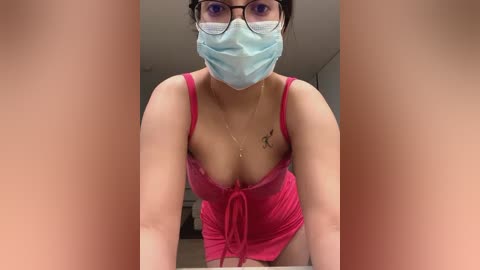 Media: Video of a woman in a pink dress with low-cut neckline, wearing a blue surgical mask and glasses, with a tattoo on her left shoulder.