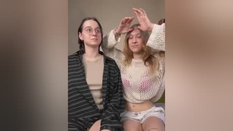 Media: Video of two young women: one in a striped shirt, the other in a knitted crop top and shorts, both with light skin, sitting indoors with a blurred background.