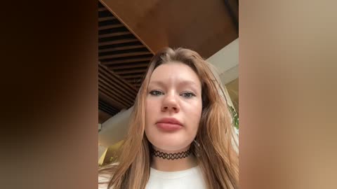 Media: Video of a young Caucasian woman with long, straight brown hair, wearing a black choker necklace, and a white shirt, standing indoors with wooden ceiling beams in the background.