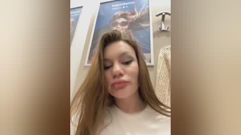 Media: A video of a young woman with long, straight brown hair, fair skin, and a slight smile. She wears a white T-shirt. Background features two posters and a beige wall.