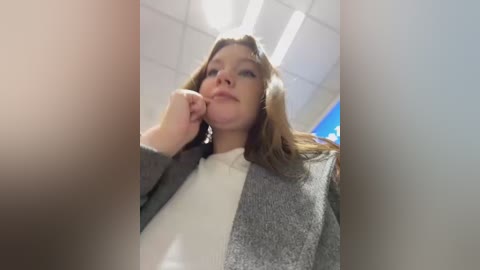 Media: Video of a Caucasian woman with shoulder-length brown hair, wearing a grey and white sweater, standing in a modern office with white ceiling tiles and bright lighting. She appears contemplative, touching her chin with her right hand.