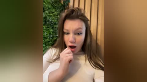 Media: Video of a young woman with long brown hair, fair skin, and a white top, biting her finger in a playful, exaggerated manner, surrounded by green foliage and wooden panels.