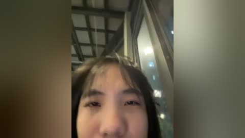 Media: Video of a young Asian woman with long black hair and pale skin, wearing a white shirt, taken from a low angle, indoors with a dimly lit, industrial ceiling.