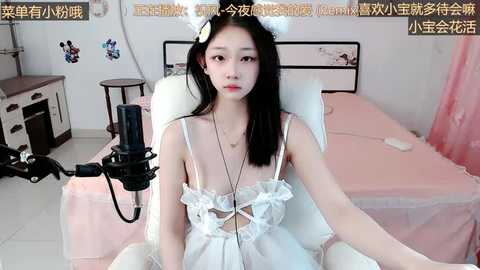 Media: Video of an East Asian woman with long black hair and white cat ears, wearing a white lace lingerie set, sitting in a pink chair, using a microphone, in a pastel-colored bedroom.