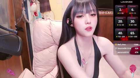 Media: Video of a slender East Asian woman with long black hair, wearing a black halter top, sitting on a beige couch in a dimly lit room.