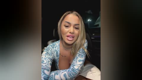 Media: Video of a light-skinned woman with long blonde hair, wearing a white, long-sleeve top with blue swirl patterns. She has a small tattoo on her chest and is seated in a car at night, looking slightly pensive.