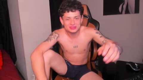 Media: Video of a shirtless young man with curly hair, tattoos, and a light tan, wearing black boxer shorts, sitting on a gaming chair in a dimly lit room.