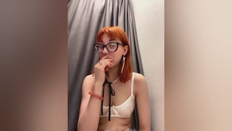 Media: Video of a fair-skinned woman with red hair, wearing glasses, a white bra, and a pearl necklace, standing in a dimly lit, grey-draped room.