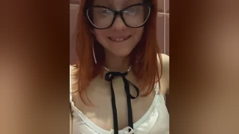 Media: A video of a young woman with long red hair, wearing black-rimmed glasses and a white blouse with a black ribbon bow, smiling.