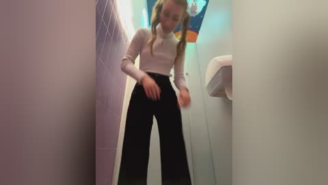 Media: Video of a young woman with light skin and long blonde hair, wearing a white long-sleeve top and black high-waisted pants, walking through a narrow, tiled hallway.