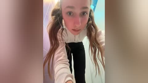 Media: A video of a young Caucasian woman with long brown hair and light skin, wearing headphones, a white sweater, and black pants, looking into the camera.