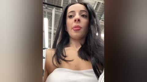 Media: A video of a young woman with long, straight black hair, fair skin, and a slender figure, wearing a white off-shoulder top, taken indoors with exposed ceiling beams and fluorescent lighting.