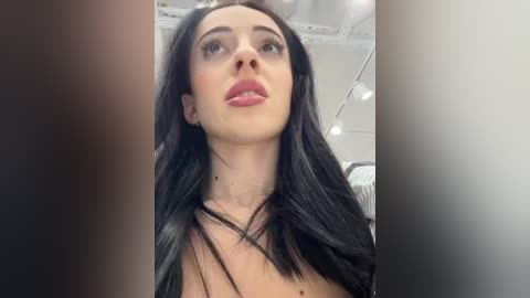 Media: Video of a young woman with long, straight black hair, fair skin, and full lips, wearing minimal makeup, captured from a low angle in an indoor setting.