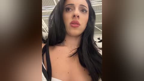 Media: Video of a fair-skinned woman with long black hair, wearing a white off-shoulder top, standing indoors with a white ceiling and blurred background.