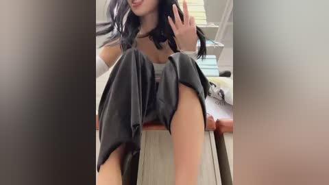 Media: Video of a woman with long black hair, fair skin, and a slender build, sitting on a chair in a brightly lit room. She wears a gray top and black pants, and raises her hand in a casual gesture.