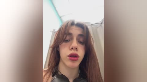 Media: Video of a young woman with long, straight brown hair, wearing a black leather jacket, and red lipstick, in a bathroom with a shower curtain.