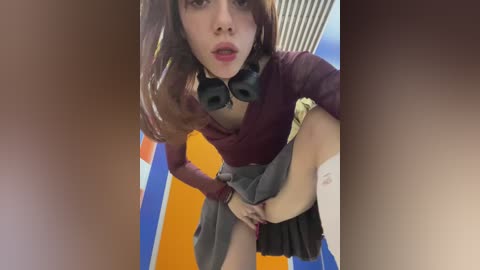 Media: A video of a young woman with fair skin and long brown hair, dressed in a maroon top and black skirt, lifting her skirt to expose her shaved pubic area. The background features blue and orange walls.