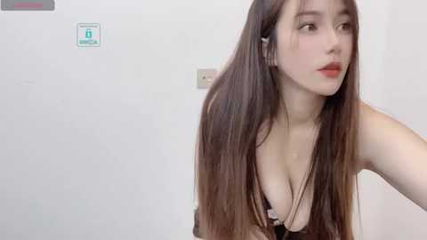 Media: Video of an East Asian woman with long brown hair, wearing a black bra, posing indoors against a plain white wall. She has fair skin, red lipstick, and a thoughtful expression.