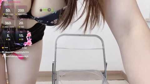 Media: Video of a light-skinned woman with long brown hair, leaning forward, wearing a black floral-patterned bra, pink panties, and a chair in a minimalist room.