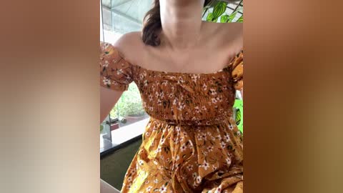 Media: Video of a woman in a brown, off-the-shoulder floral dress with a gathered waist, standing indoors near a window with green plants.