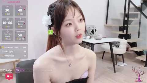 Media: Video of an East Asian woman with light skin and long brown hair, wearing a black strapless top, sitting in a modern, minimalist room with a staircase and white table.