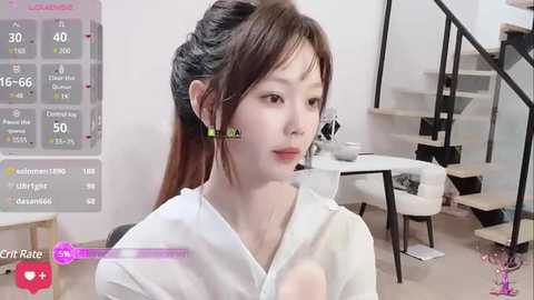 Media: Video of an East Asian woman with straight, shoulder-length brown hair, wearing a white top, seated indoors. Background includes a staircase and modern furniture.